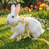 Load image into Gallery viewer, A white bunny walking in a sunny flower garden wearing a cute yellow rabbit harness featuring an adorable plush animal charm.
